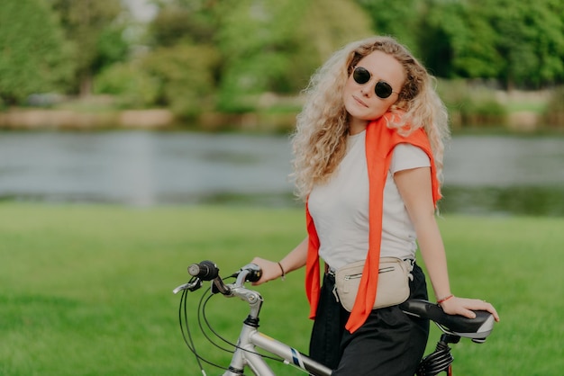 Une jolie femme fait du vélo a des cheveux blonds touffus se tient près de son vélo porte des lunettes de soleil et un t-shirt blanc aime voyager à l'air frais petite rivière ou lac en arrière-plan herbe verte