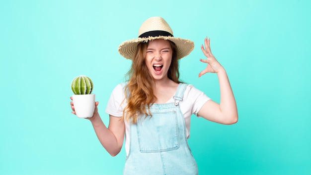 Jolie femme fermière à tête rouge criant avec les mains en l'air et tenant un cactus