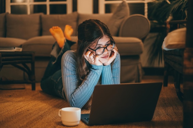 Une jolie femme gaie et jeune portant des lunettes à l'aide d'un ordinateur sur le tapis à la maison la nuit