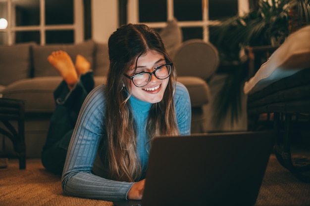 Une jolie femme gaie et jeune portant des lunettes à l'aide d'un ordinateur sur le tapis à la maison la nuit