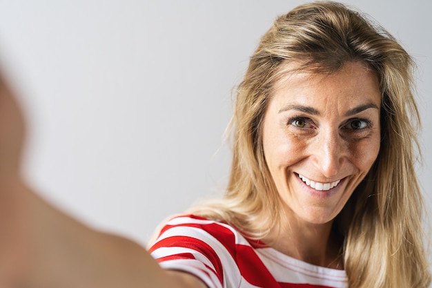 Jolie femme habillée avec désinvolture et souriant largement. Belle femme ayant une expression joyeuse tout en posant sur fond blanc, étirant le bras vers la caméra pour un selfie