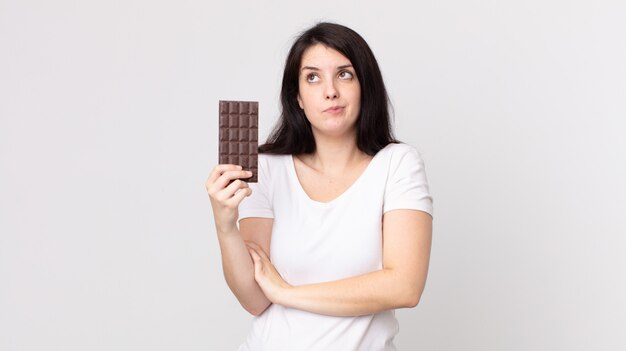 Jolie femme haussant les épaules, se sentant confuse et incertaine et tenant une barre de chocolat