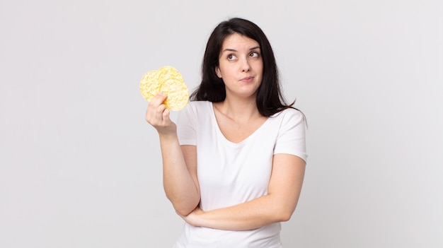 Jolie femme haussant les épaules, se sentant confuse et incertaine et tenant un régime de gâteaux de riz