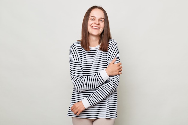 Jolie femme heureuse souriante aux cheveux bruns portant une chemise rayée debout isolée sur fond gris regardant la caméra en riant exprimant des émotions positives