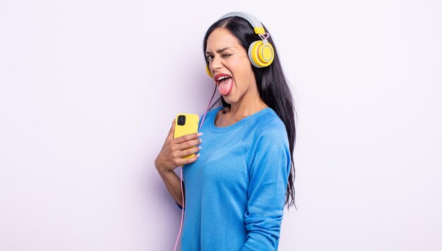 Jolie femme hispanique avec une attitude joyeuse et rebelle, plaisantant et tirant la langue. concept de smartphone et casque