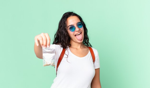 Jolie femme hispanique avec une attitude joyeuse et rebelle, plaisantant et tirant la langue et tenant un sac de marihuana