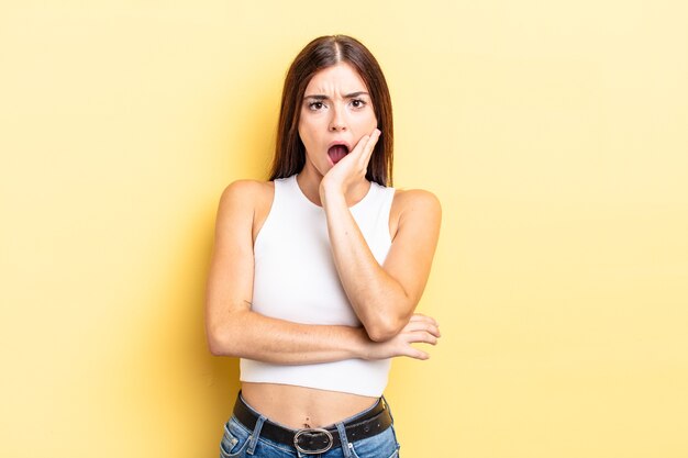 Jolie femme hispanique bouche bée sous le choc et l'incrédulité, la main sur la joue et le bras croisés, se sentant stupéfaite et étonnée