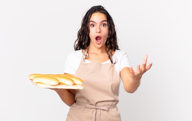 Jolie femme hispanique chef étonnée, choquée et étonnée d'une incroyable surprise et tenant un troy de petits pains