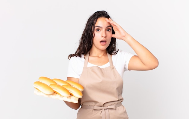 Jolie femme hispanique chef à la recherche de bonheur, d'étonnement et de surprise et tenant un troy de petits pains