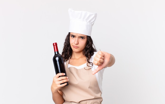 Jolie femme hispanique chef se sentant croisée, montrant les pouces vers le bas et tenant une bouteille de vin