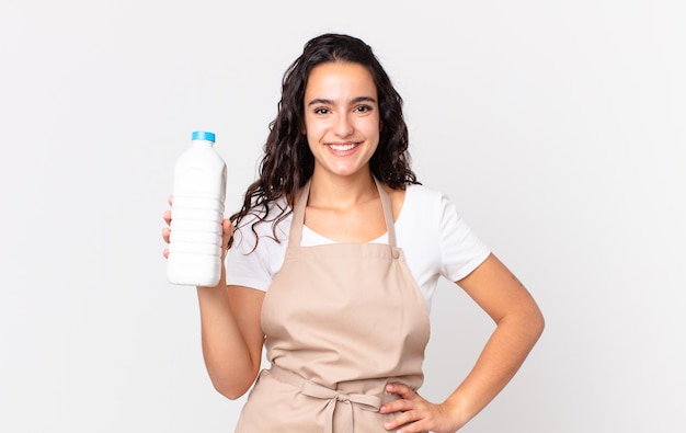 jolie femme hispanique chef souriante joyeusement avec une main sur la hanche et confiante et tenant une bouteille de lait