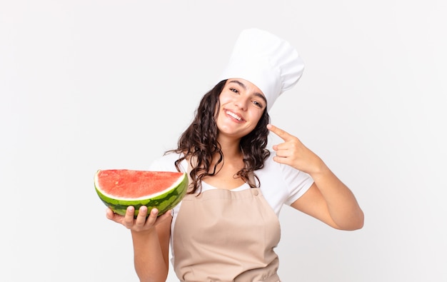 Jolie femme hispanique chef souriante pointant avec confiance vers son propre large sourire et tenant une pastèque