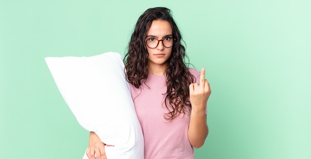 Jolie femme hispanique se sentant en colère, agacée, rebelle et agressive et portant un pyjama avec un oreiller