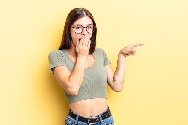 Jolie femme hispanique se sentant heureuse, choquée et surprise, couvrant la bouche avec la main et pointant vers l'espace de copie latéral