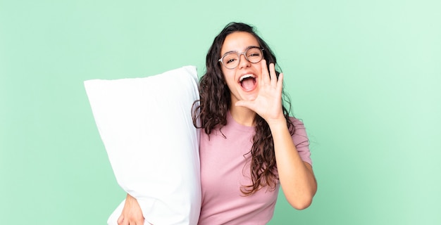 jolie femme hispanique se sentant heureuse, donnant un grand cri avec les mains à côté de la bouche et portant un pyjama avec un oreiller