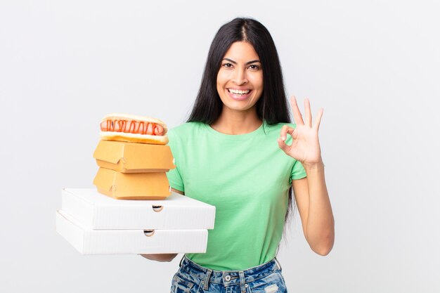 Jolie femme hispanique se sentant heureuse, montrant son approbation avec un geste correct et tenant des boîtes de restauration rapide à emporter