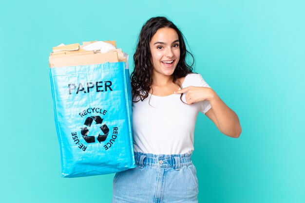 Jolie femme hispanique se sentant heureuse et se montrant elle-même avec un excité et tenant un sac en papier recyclé