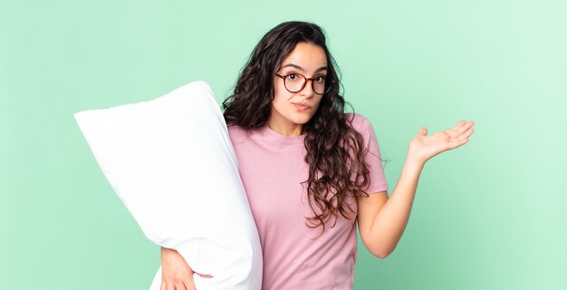 Jolie femme hispanique se sentant perplexe et confuse et doutant et portant un pyjama avec un oreiller