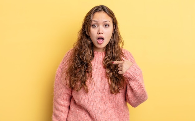 Jolie femme hispanique semblant choquée et surprise avec la bouche grande ouverte, pointant vers soi