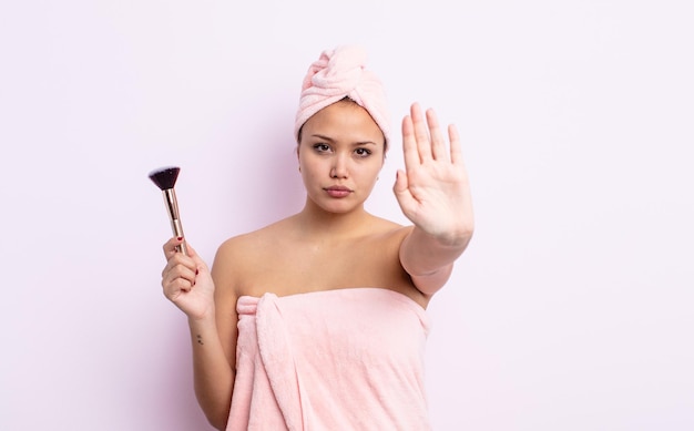 Jolie femme hispanique semblant sérieuse montrant la paume ouverte faisant un geste d'arrêt. concept de pinceau de maquillage