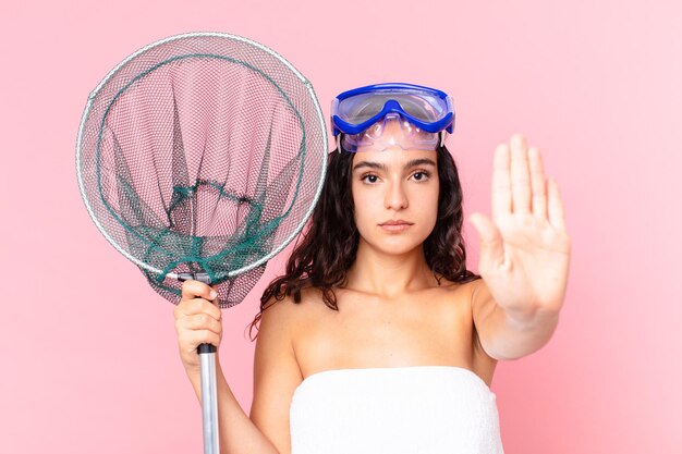 Jolie femme hispanique à la sérieuse montrant la paume ouverte faisant un geste d'arrêt avec des lunettes et un filet de pêche