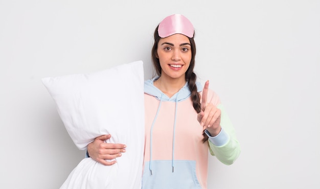 Jolie femme hispanique souriant fièrement et en toute confiance faisant le numéro un. concept pyjama et oreiller