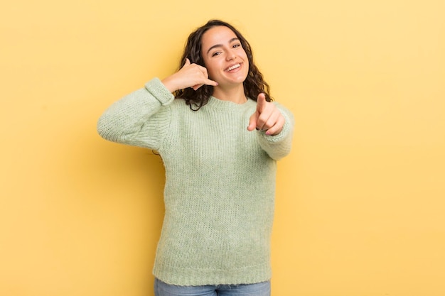 Jolie femme hispanique souriant joyeusement et pointant vers la caméra tout en vous appelant plus tard en parlant au téléphone