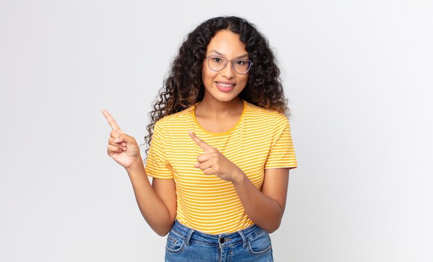 jolie femme hispanique souriant joyeusement et pointant vers le côté et vers le haut avec les deux mains montrant l'objet dans l'espace de copie