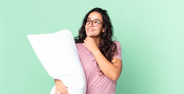 Jolie femme hispanique souriant joyeusement et rêvant ou doutant et portant un pyjama avec un oreiller