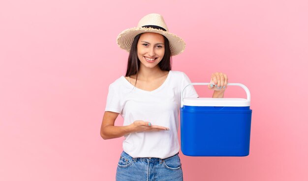 Jolie femme hispanique souriant joyeusement, se sentant heureuse et montrant un concept et tenant un réfrigérateur portable