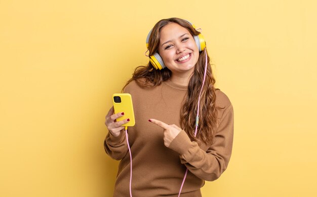 Jolie femme hispanique souriant joyeusement se sentant heureuse et pointant vers le côté casque et concept de smartphone