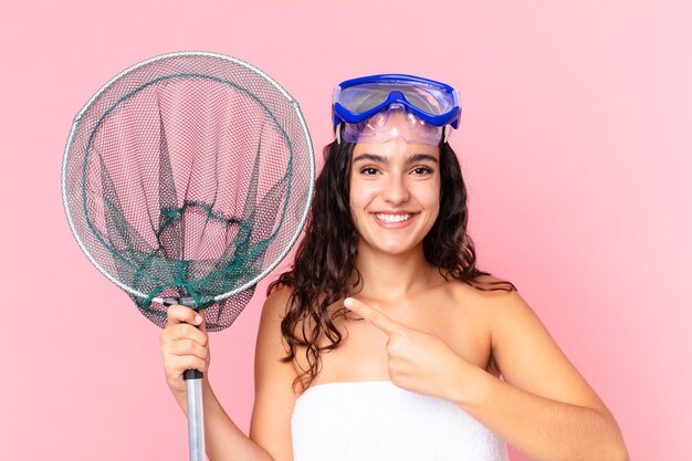 Jolie femme hispanique souriant joyeusement, se sentant heureuse et pointant vers le côté avec des lunettes et un filet de pêche