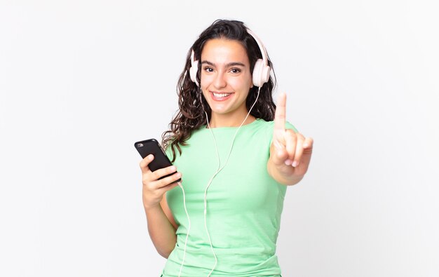 Jolie femme hispanique souriante fièrement et avec confiance faisant le numéro un avec des écouteurs et un smartphone