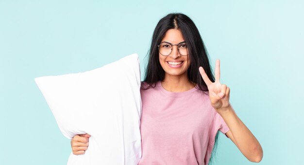 Jolie femme hispanique souriante et semblant heureuse, gesticulant la victoire ou la paix et portant un pyjama avec un oreiller