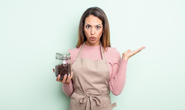 Jolie femme hispanique à la surprise et choquée avec la mâchoire tombée tenant un concept de grains de café d'objet