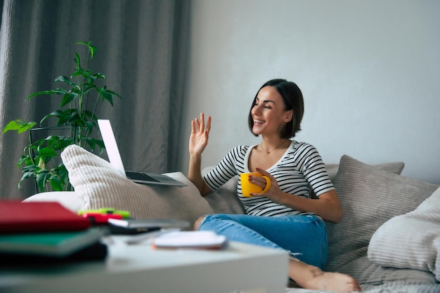 Jolie femme indépendante souriante travaillant sur un ordinateur portable et ayant une conversation en ligne alors qu'elle est assise sur le grand canapé et boit du café à la maison