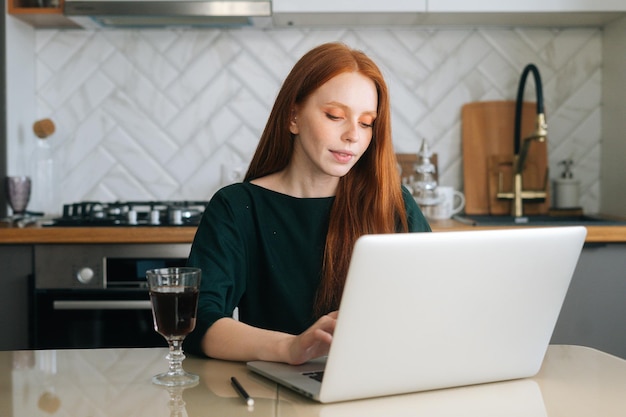 Jolie femme indépendante travaillant en tapant sur un ordinateur portable assis à table avec un verre de boisson alcoolisée dans la cuisine