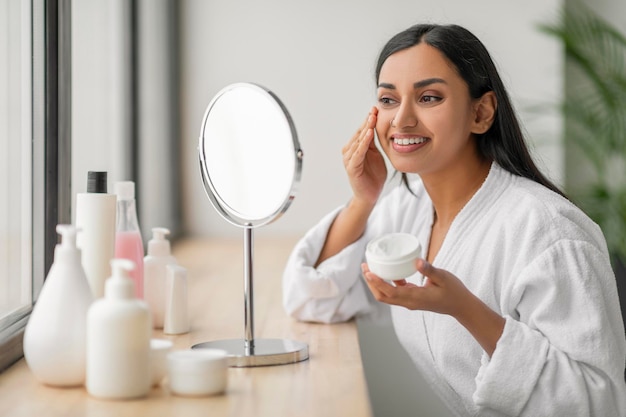 Jolie femme indienne millénaire appliquant une crème nourrissante sur le visage