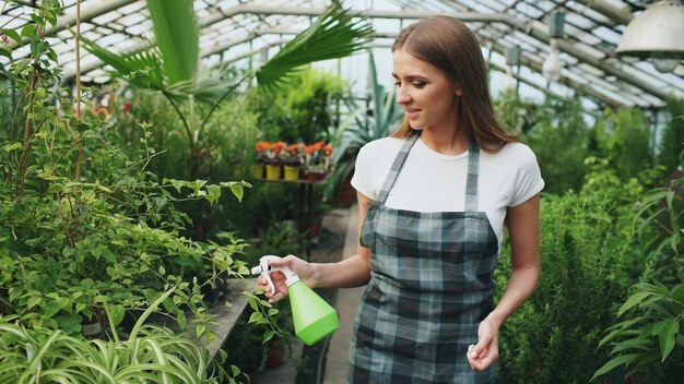 Jolie femme jardinier en tablier arrosant les plantes et les fleurs avec un pulvérisateur de jardin en serre