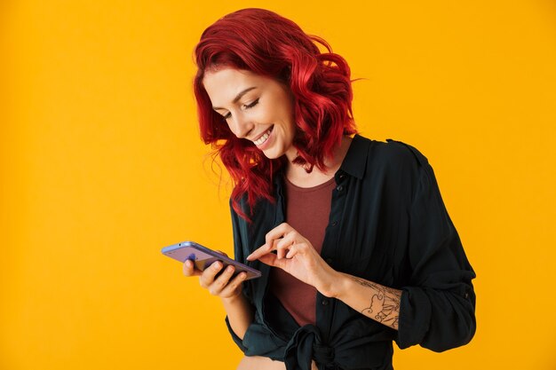 jolie femme joyeuse aux cheveux roux bouclés souriant et tapant sur téléphone portable isolé