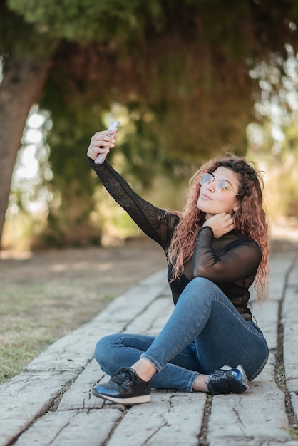 Jolie femme latine avec des lunettes faisant un autoportrait avec son smarphone assis sur un chemin en bois dans le domaine à l'extérieur