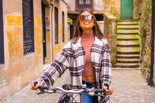 Jolie femme latine avec des lunettes de soleil faisant du tourisme à vélo dans la ville à travers la vieille ville Écotourisme au printemps en vacances modèle souriant