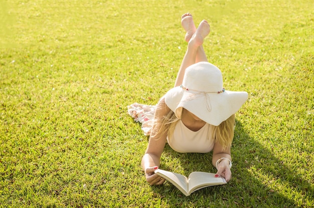 Jolie femme lisant un livre allongé sur la pelouse, vu d'en haut avec un chapeau d'été.