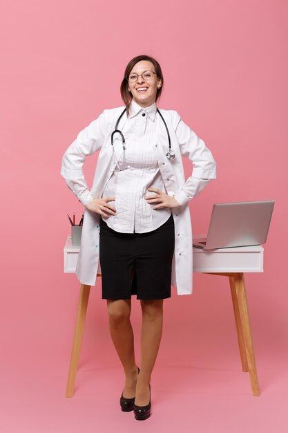 Une jolie femme médecin se tient devant le bureau avec un ordinateur pc, un document médical à l'hôpital isolé sur fond de mur rose pastel. Femme en robe médicale lunettes stéthoscope. Concept de médecine de santé