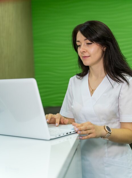 Jolie femme médecin travaillant sur ordinateur portable au bureau. Médecin brune en gommage blanc.