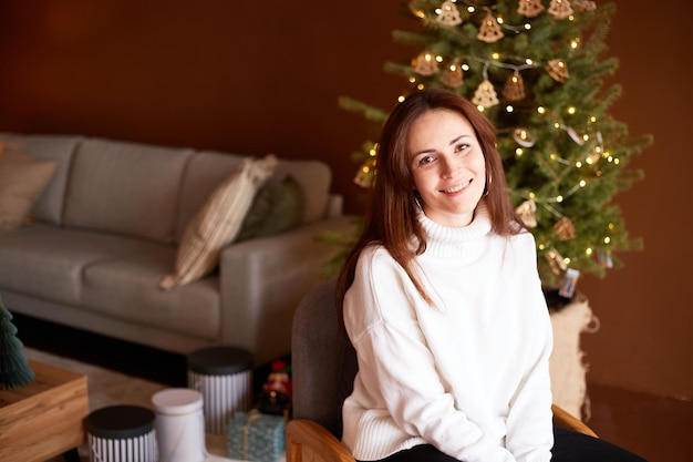 Jolie femme millénaire assise sur une chaise dans le salon décoré Arbre de Noël avec décoration sur fond