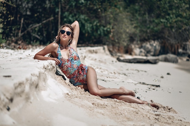 Jolie femme mince en lunettes de soleil et maillot de bain sur la plage blanche contre les arbres, bras dans le haer ; notion de bronzage.