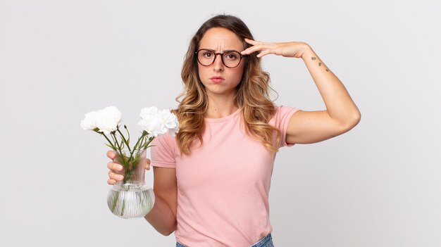 Jolie femme mince se sentant confuse et perplexe montrant que vous êtes fou et tenant des fleurs décoratives