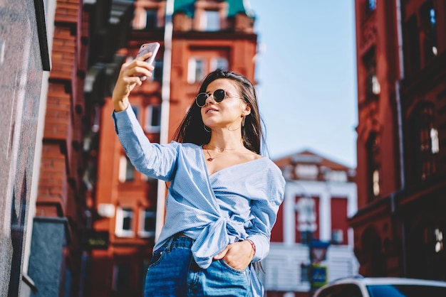 Jolie femme à la mode dans des lunettes de soleil tenant un téléphone portable à la main en regardant la caméra du smartphone