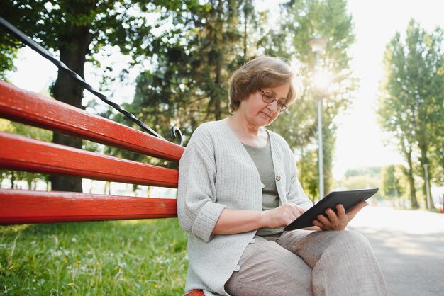 Jolie femme mûre assise sur un banc tenant et utilisant une tablette numérique dans le parc le jour d'été